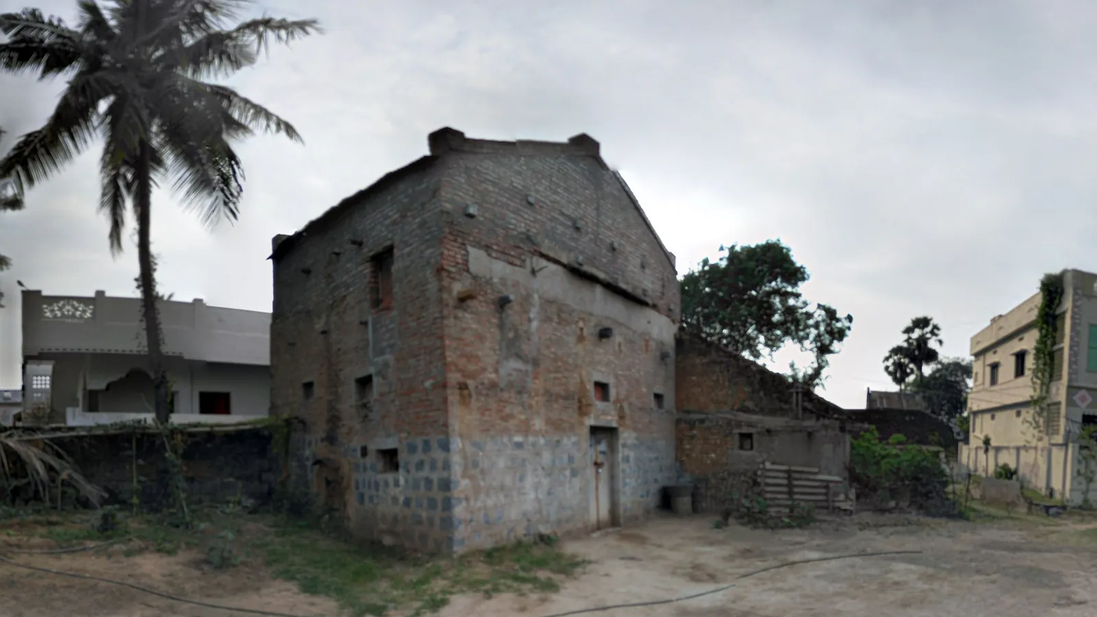 Tall square brick flue-curing barn with tie-rod anchor plates and small vents, a low furnace shed attached on the right, standing on a sandy village yard under an overcast sky with a coconut palm on the left.