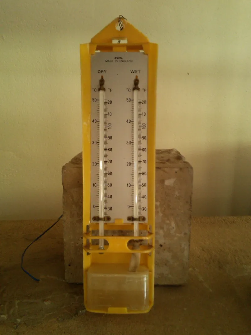 A yellow plastic wall-mounted dry-and-wet-bulb hygrometer (Zeal, made in England) with two glass thermometers marked DRY and WET and a water reservoir with a wick, standing against a cream wall.