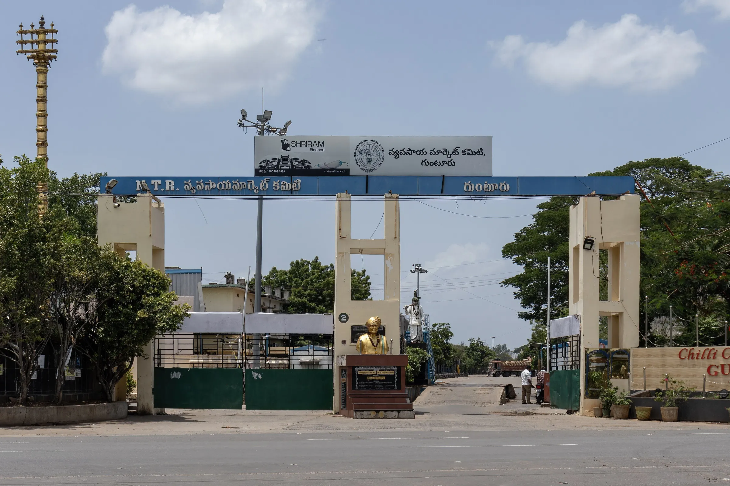 The right-hand side of the Guntur agricultural market-yard entrance: a cream gate pillar under a blue beam, the road leading into the yard and a lorry in the distance.